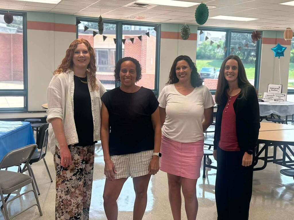Four district staff members are standing together in a school cafeteria decorated for a kindergarten event, posing for a group photo.