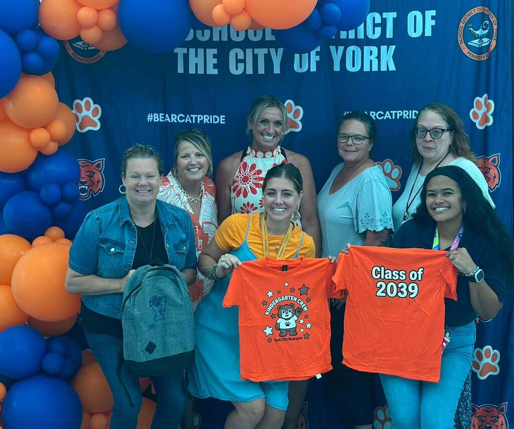 A group of district staff members are posing with a kindergarten backpack and Kindergarten Crew shirts in front of a Bearcat Pride backdrop decorated with blue and orange balloons.