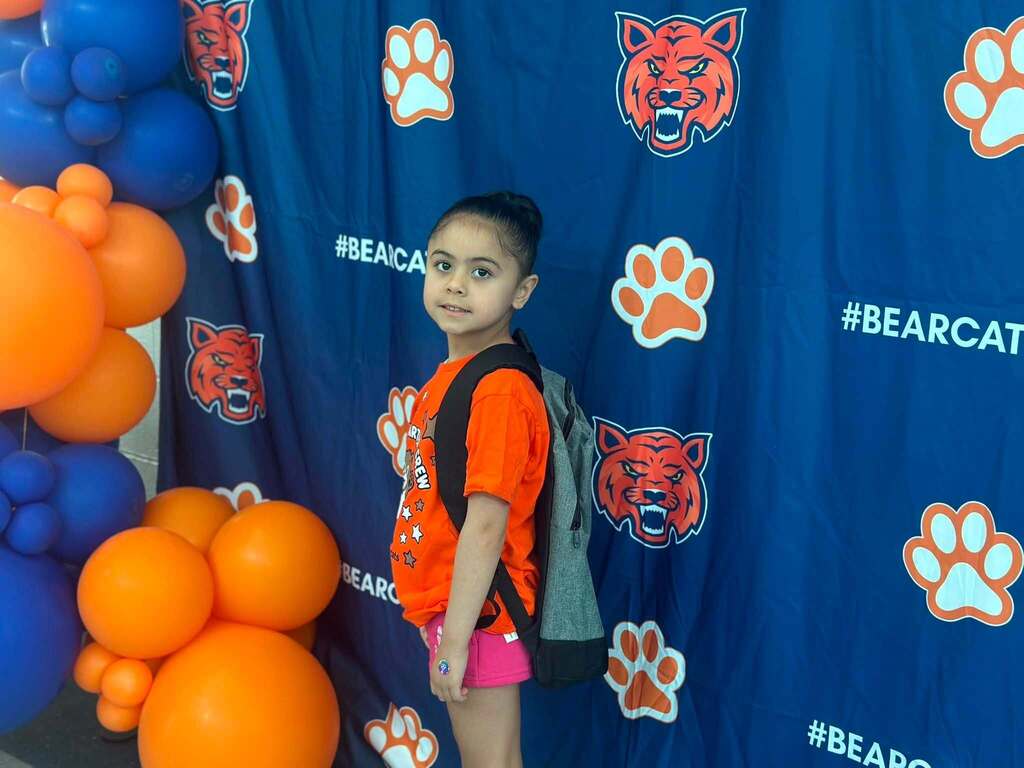 A young student wearing an orange Kindergarten Crew shirt and backpack is posing in front of a Bearcat Pride backdrop with blue and orange balloon decorations.