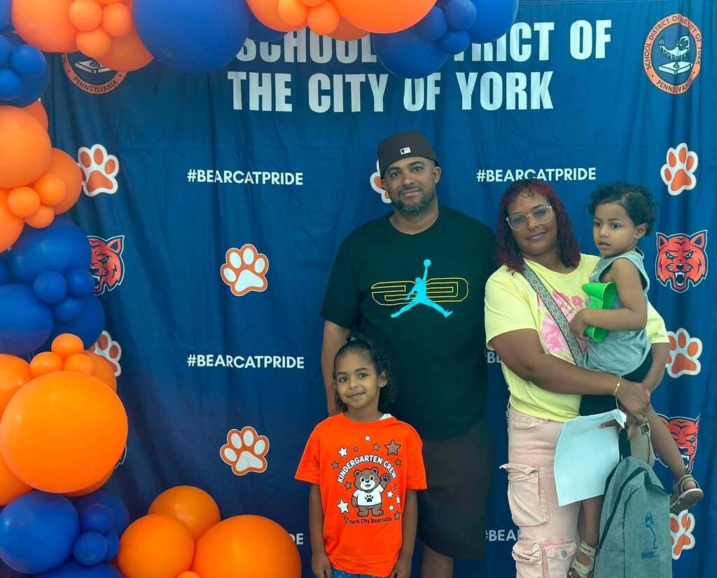 A district family consisting of an adult man and woman and two young children, are posing together at kindergarten orientation in front of a School District of the City of York Bearcat Pride backdrop, with their future kindergartener wearing an orange Kindergarten Crew shirt.
