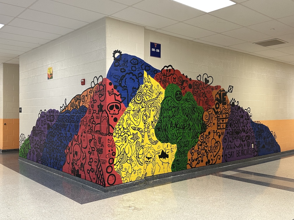 A colorful hand-drawn mural wraps around a hallway wall inside a school, featuring vibrant sections of blue, red, yellow, green, orange, and purple filled with creative doodles, symbols, and school spirit artwork.