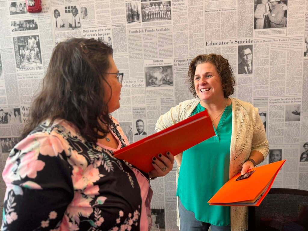 Two district staff members smile and talk while holding orange folders in front of a wall covered with historic newspaper pages.
