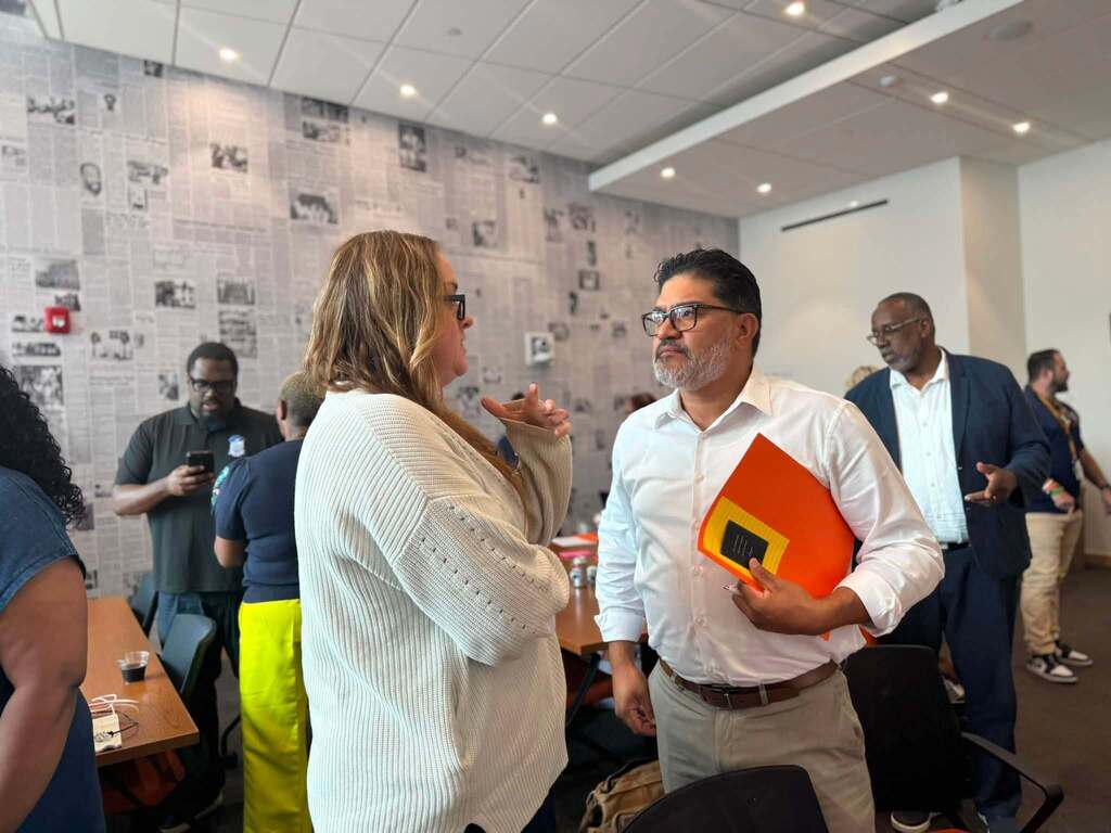 Two district staff members talk during a meeting while one holds an orange folder, with other attendees networking in the background.