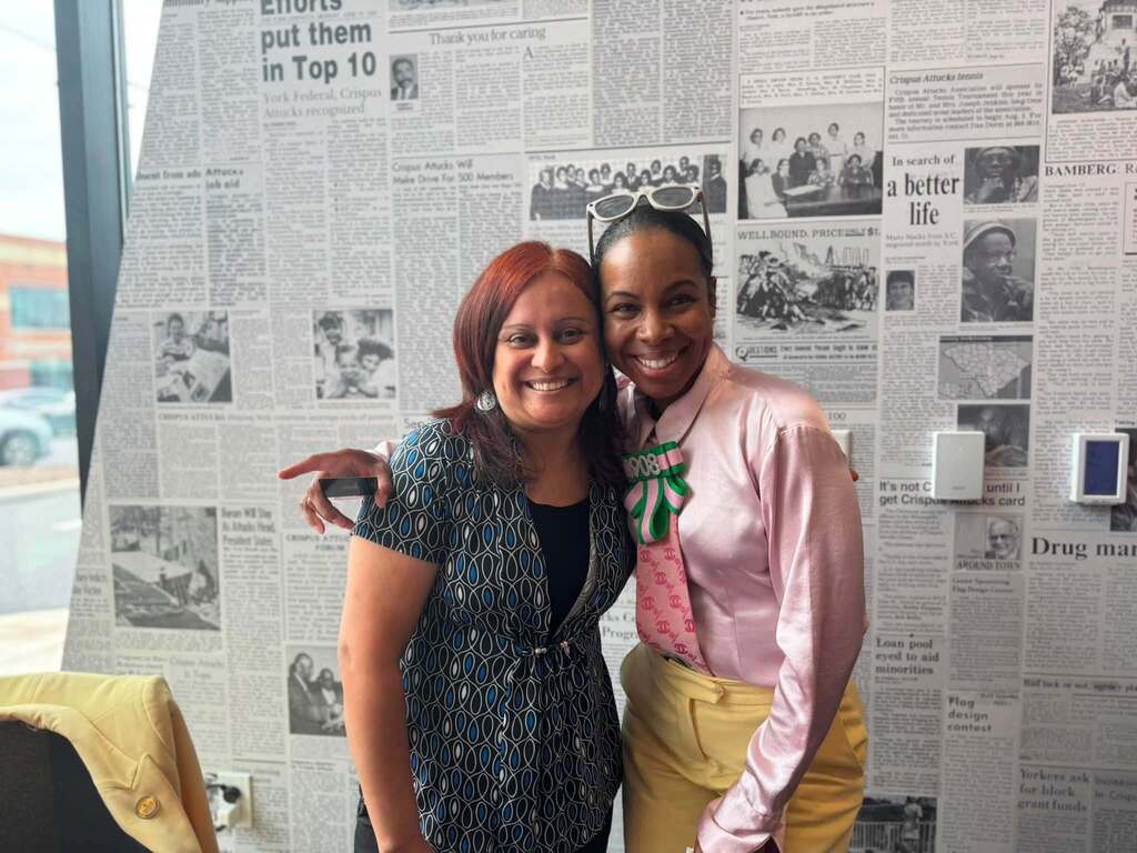 Two smiling district staff members pose with their arms around each other in front of a wall covered with historic newspaper pages.
