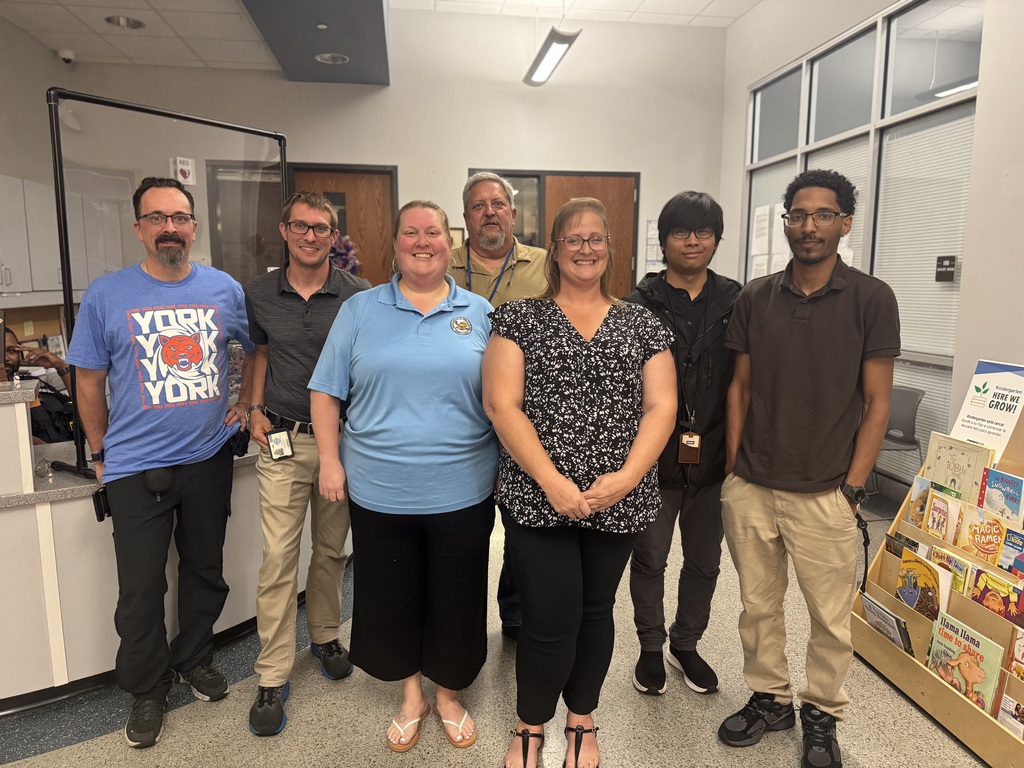The district's Information and Technology Department are smiling for a group photo in a lobby area, with desks, windows, and a colorful children’s book display behind them.