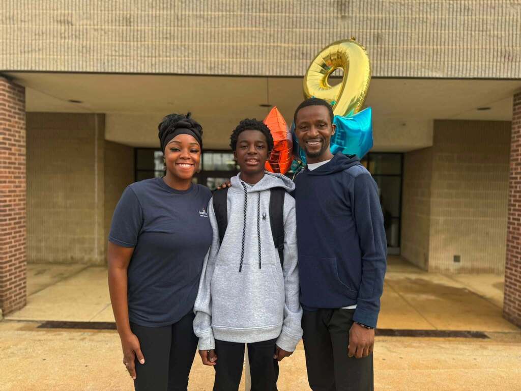 A student is standing in the middle of an adult man and woman outside a school with colorful balloons.