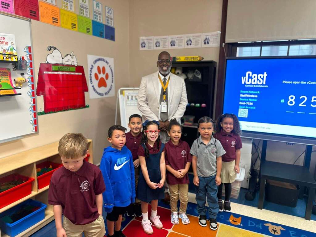 A group of students are posing with a district staff member in an elementary classroom.