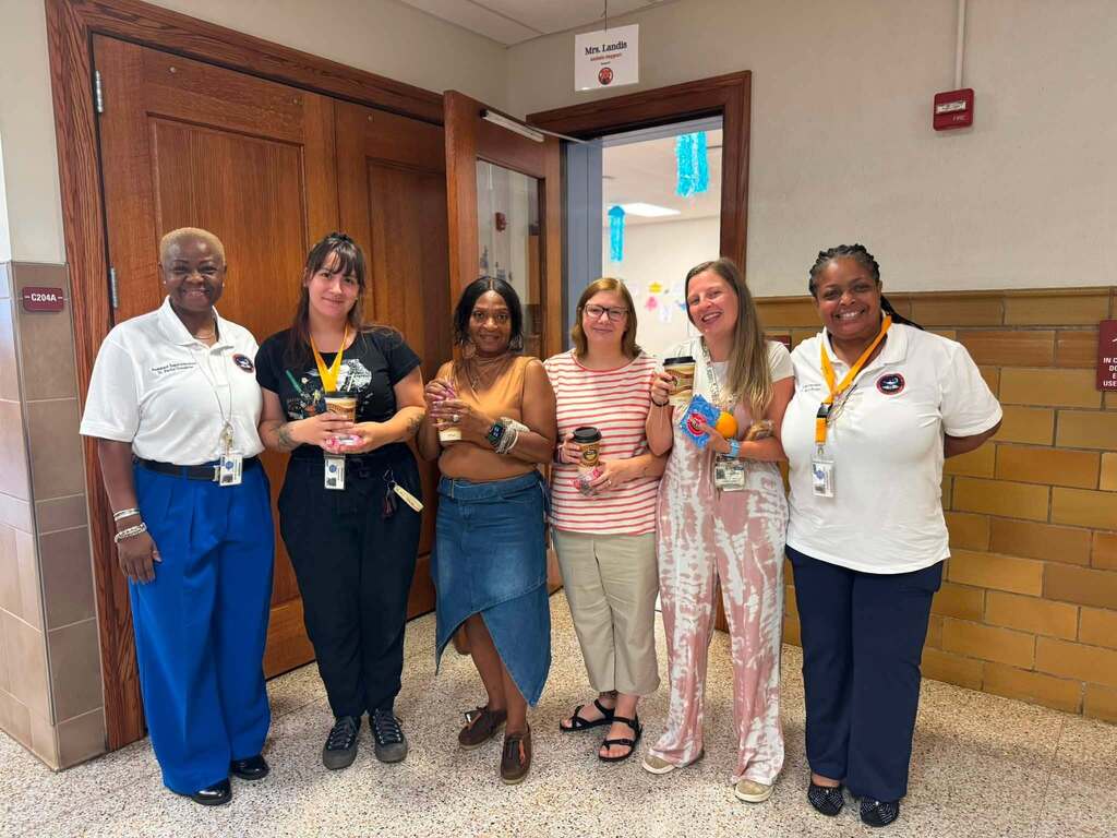 District Superintendent and Assistant Superintendent are posing with district staff members smile for a photo while holding coffee cups in a school hallway.