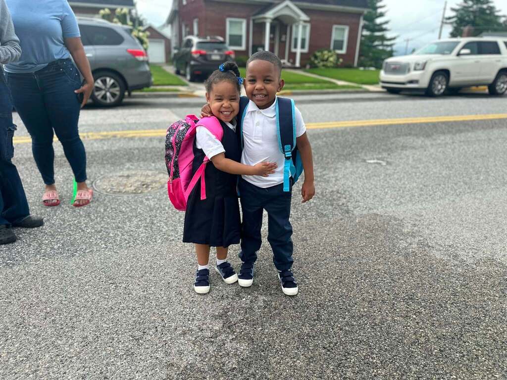 Two smiling students with backpacks are hugging each other while standing outside on a street.