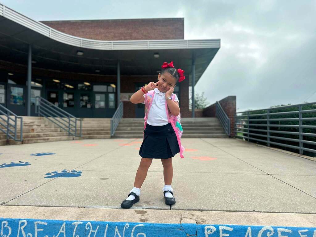 A young student is posing outside a school entrance with a pink backpack and red hair bows.