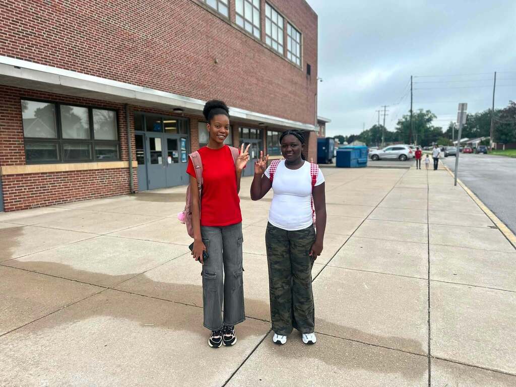 Two students are posing outside a school building, smiling and making peace signs.