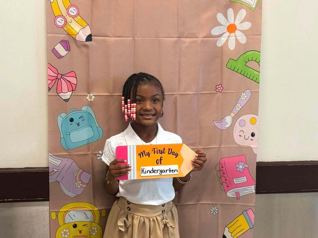 A smiling student is holding a “My First Day of Kindergarten” sign in front of a colorful school-themed backdrop.