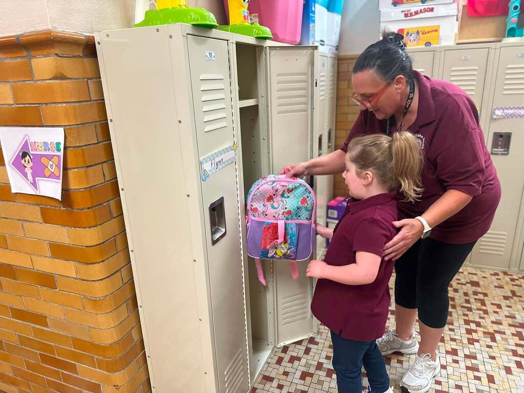 A student is putting away their backpack in a school locker with help from a district staff member.