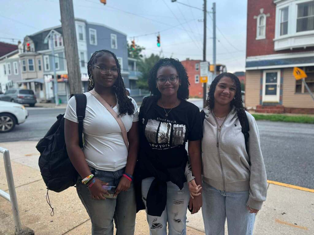 Three students with backpacks are posing together on a sidewalk outside a school.