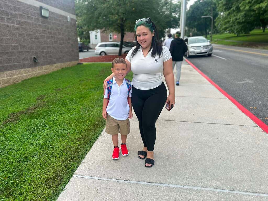 A student with a backpack is standing next to an adult on a sidewalk outside a school. Other people can be seen in the background.