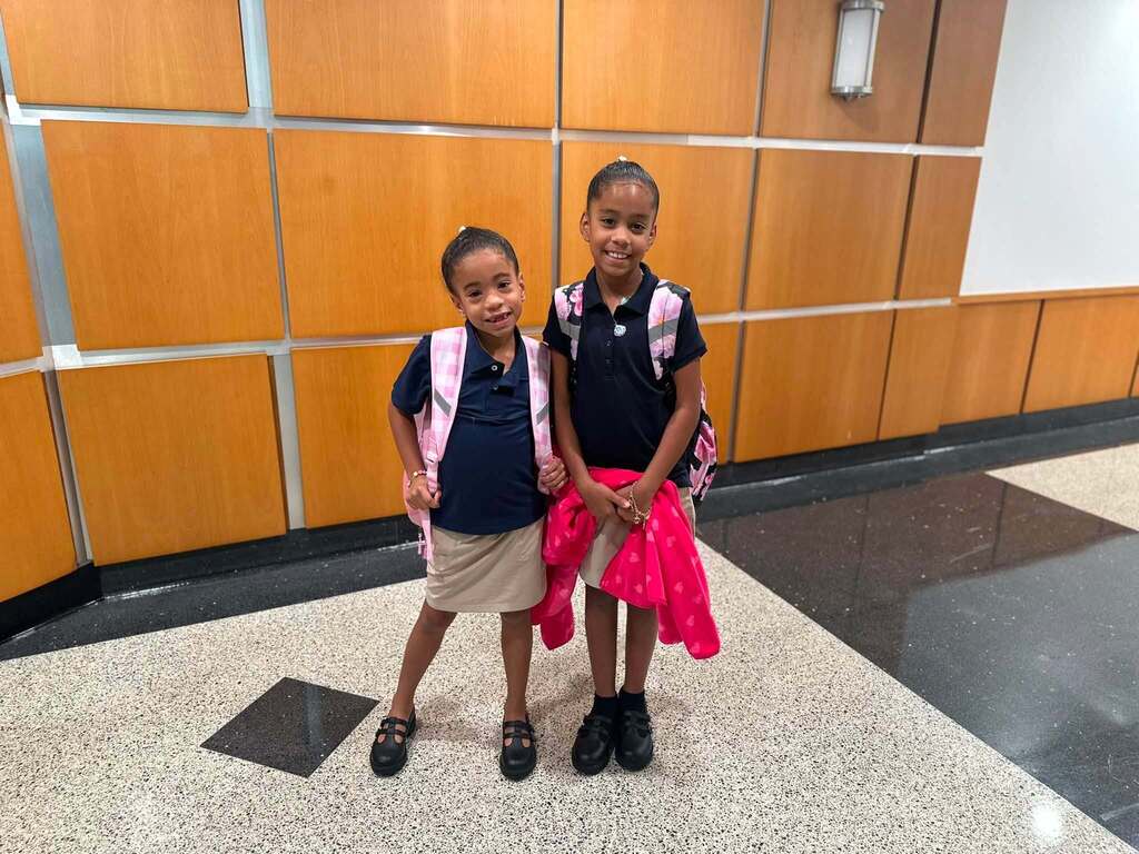 Two students are posing for a photo inside a school building, wearing uniforms and backpacks.