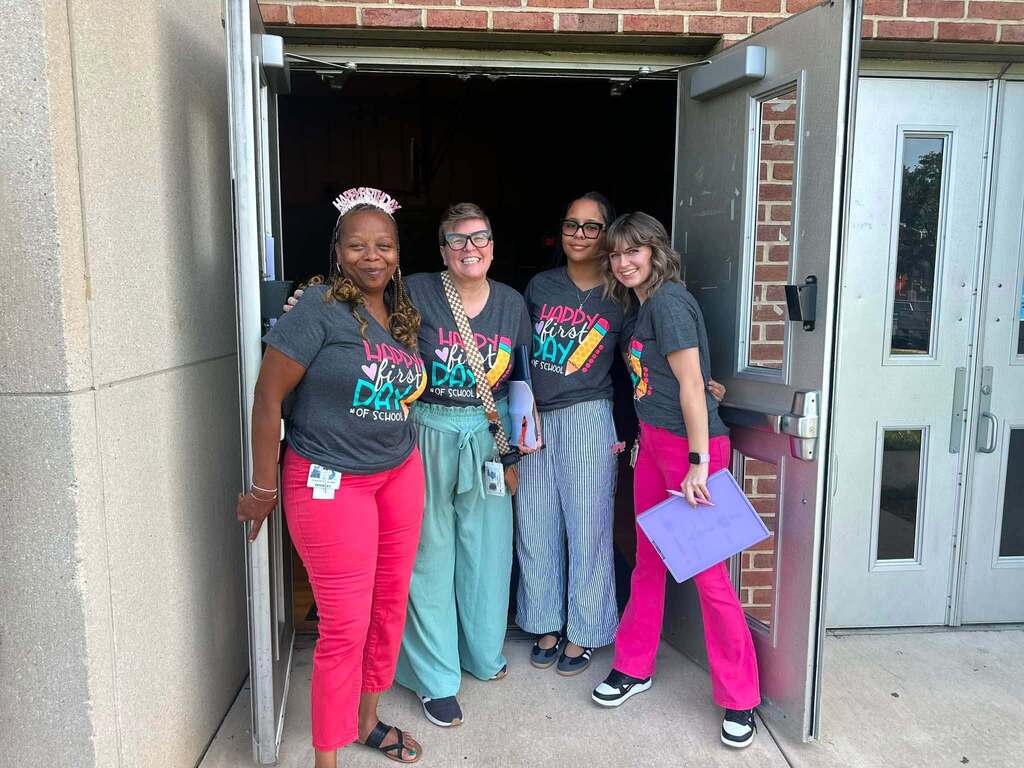 Four district staff members are posing together in a doorway, wearing matching “Happy First Day of School” shirts.