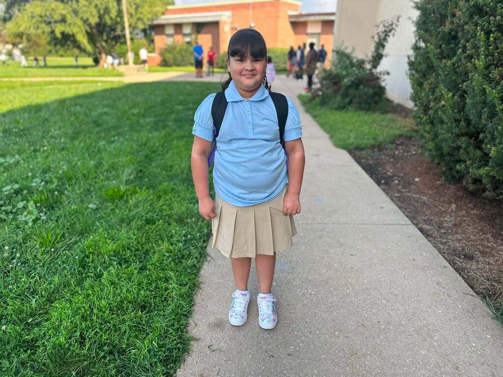 A student in a school uniform is posing on a sidewalk with a backpack.