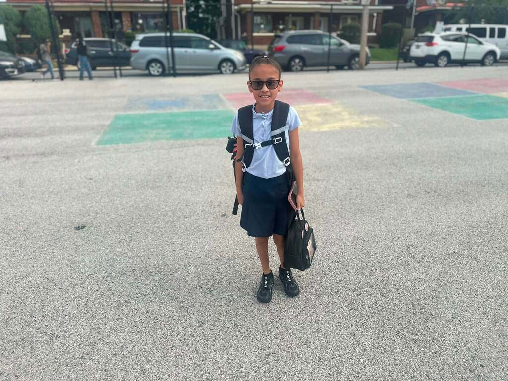 A student is posing outdoors wearing a school uniform, backpack, and glasses.