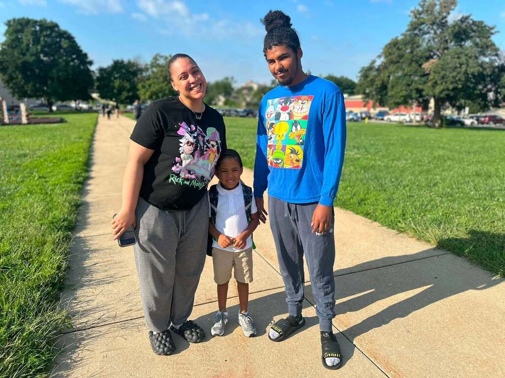 Two adults and a young child are posing for a photo on a sunny walkway surrounded by green grass.