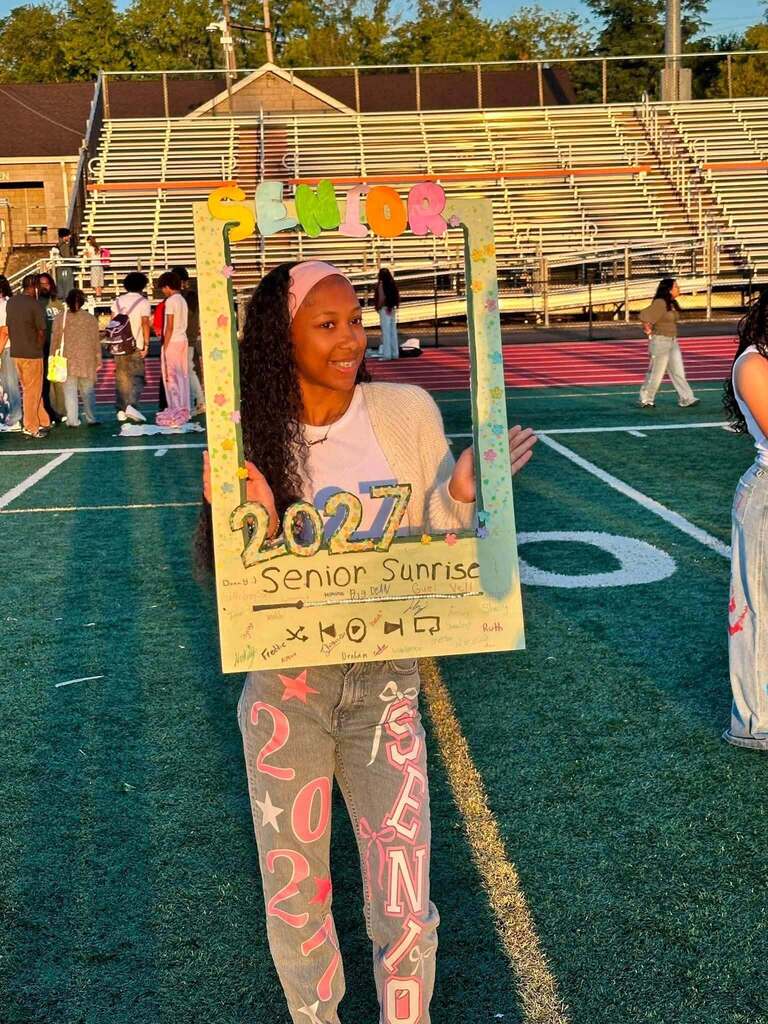 A student is posing on a football field holding a decorated Senior Sunrise 2027 frame.