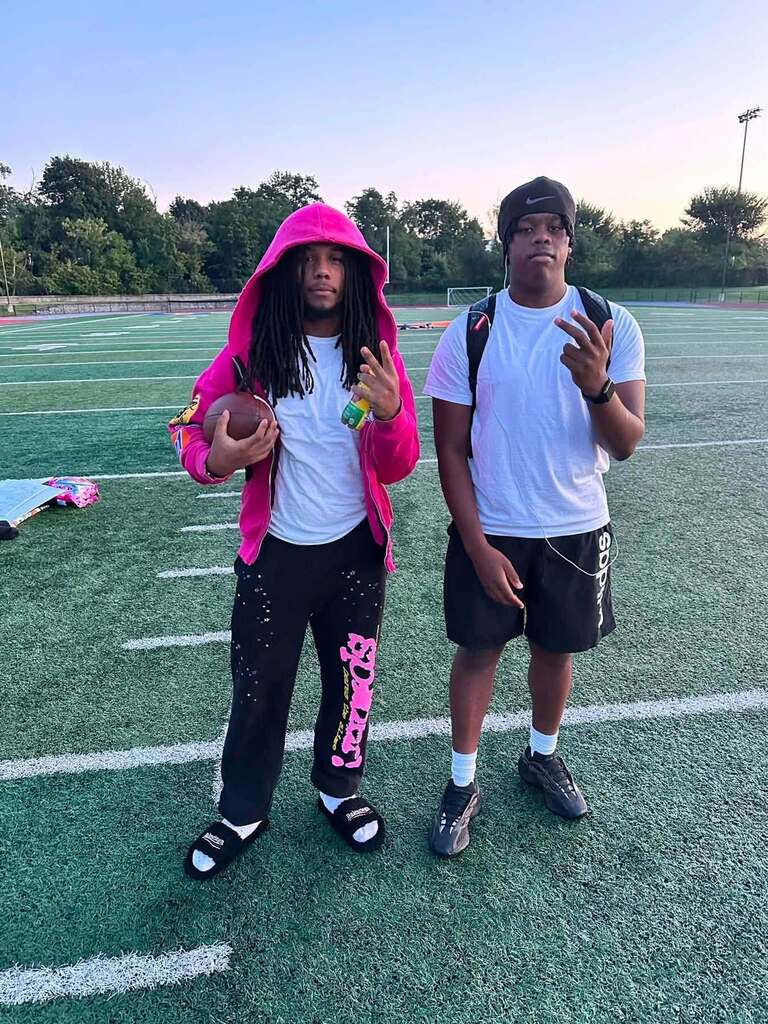 Two students are posing together on a football field, with one holding a football and wearing a bright pink hoodie.