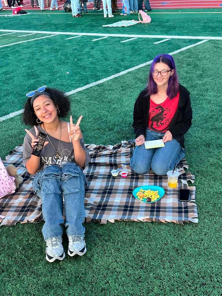 Two students are sitting on a picnic blanket on a football field, smiling and posing with peace signs.