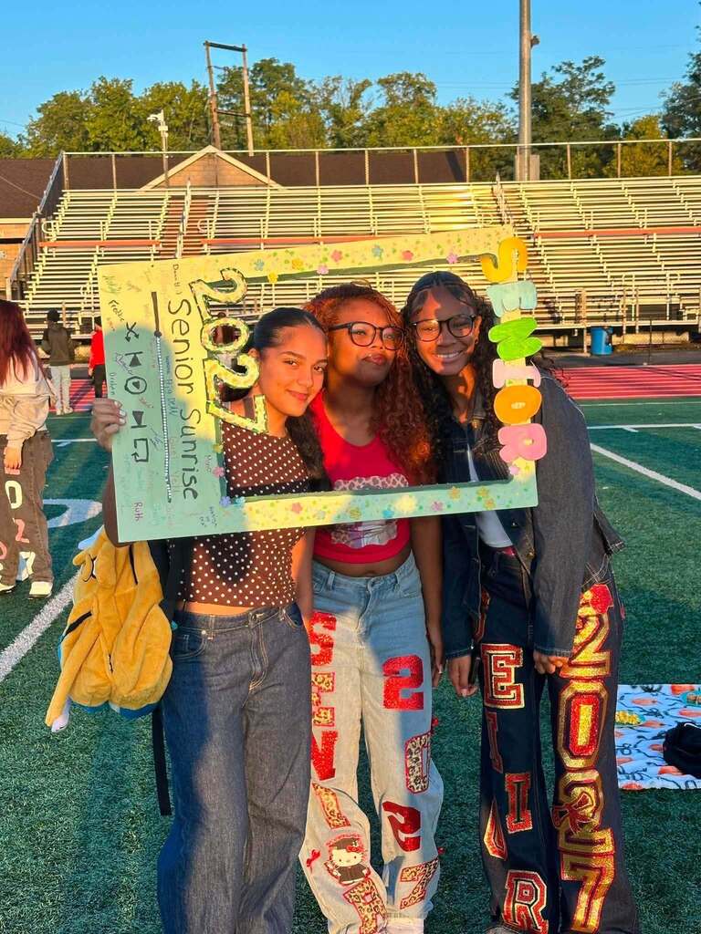 Three students are posing together on a football field with a decorated Senior Sunrise frame.