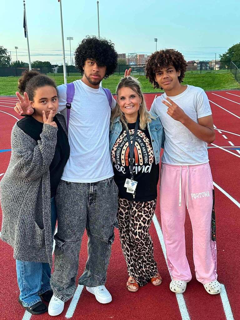 Three students and a district staff member are posing together on a track, smiling and making peace signs.