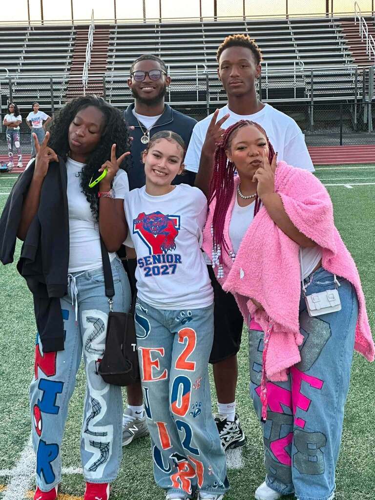 Five students are posing together on a football field, smiling and wearing colorful Senior 2027 outfits.