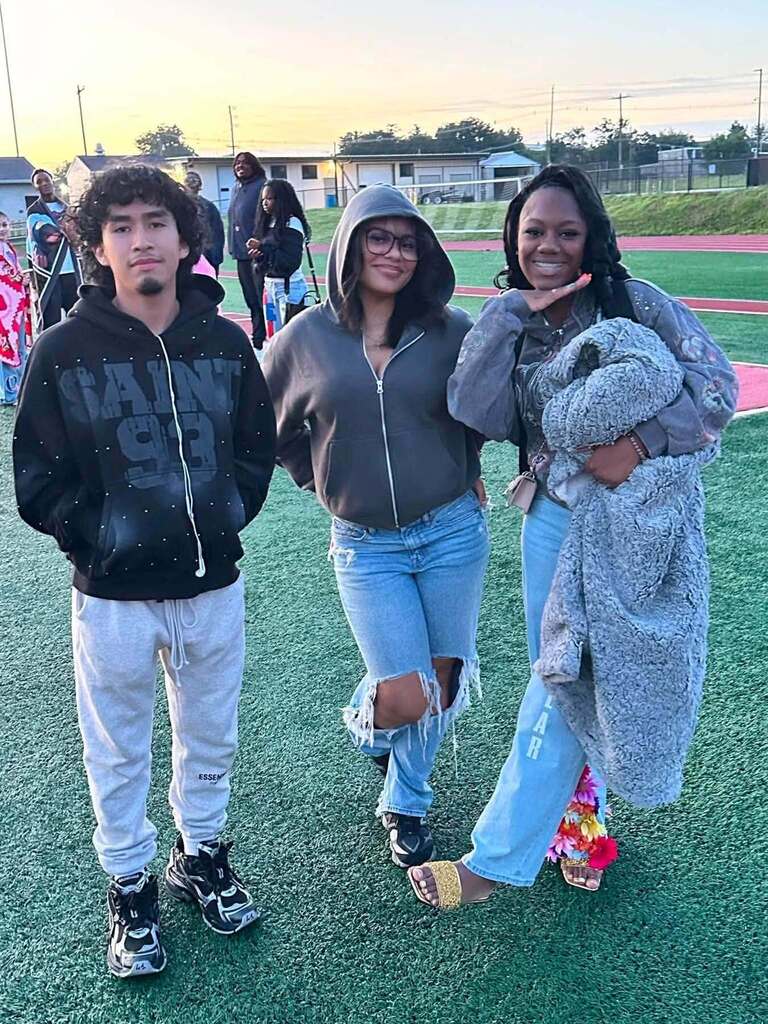 Three students are posing together on a football field during sunrise.
