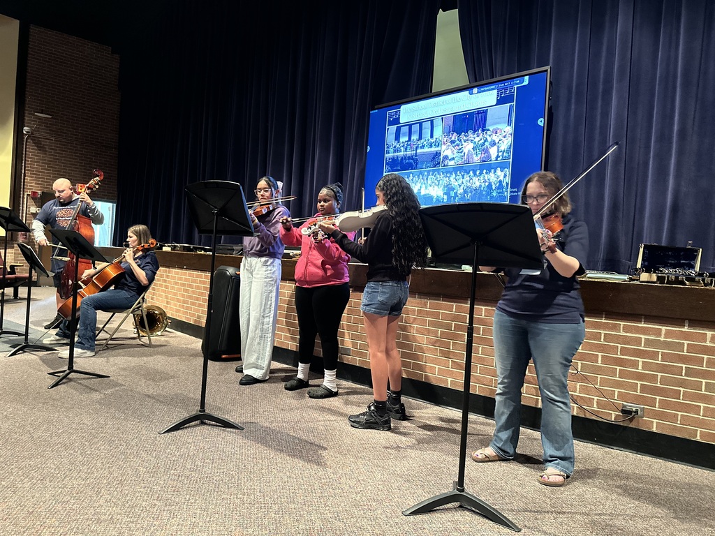District staff members and students are performing with string instruments in a school auditorium.