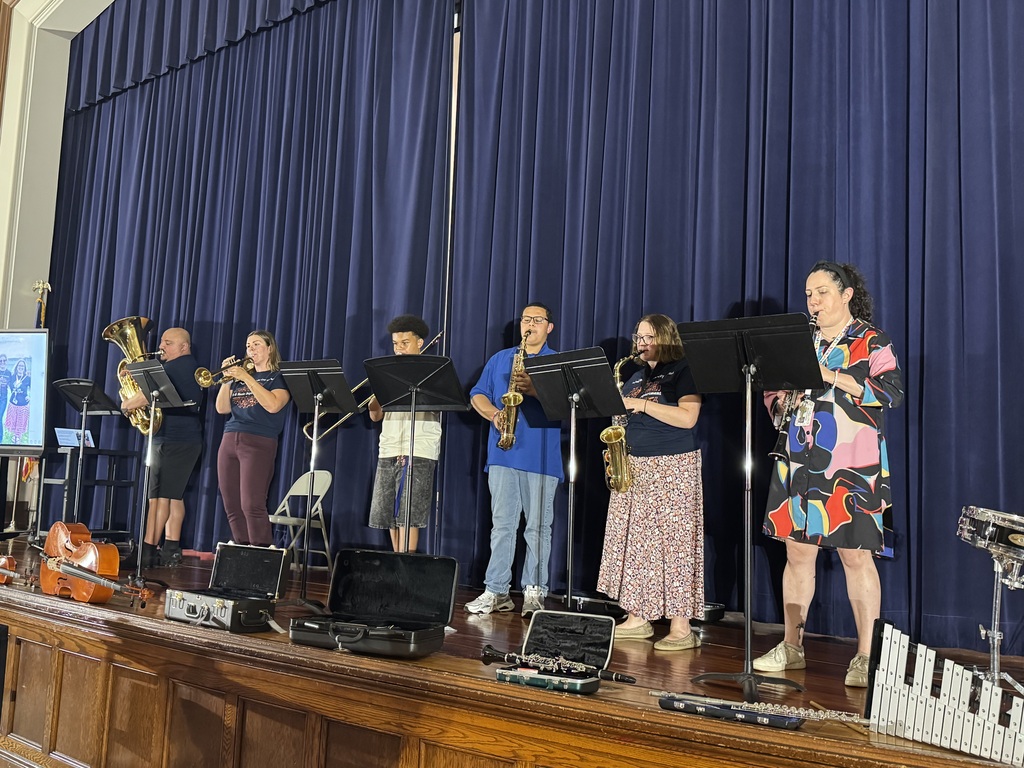 District staff members and students are performing on stage in front of blue curtains, playing brass and woodwind instruments.