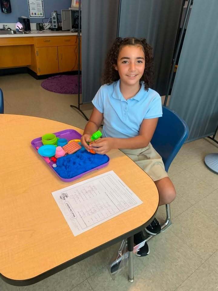 A student is working with blue sensory sand and colorful tools at a classroom table.