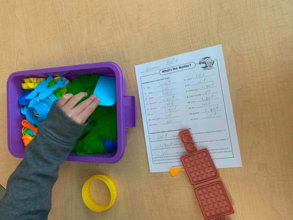 A student is exploring green sensory sand with colorful tools beside a worksheet about states of matter.