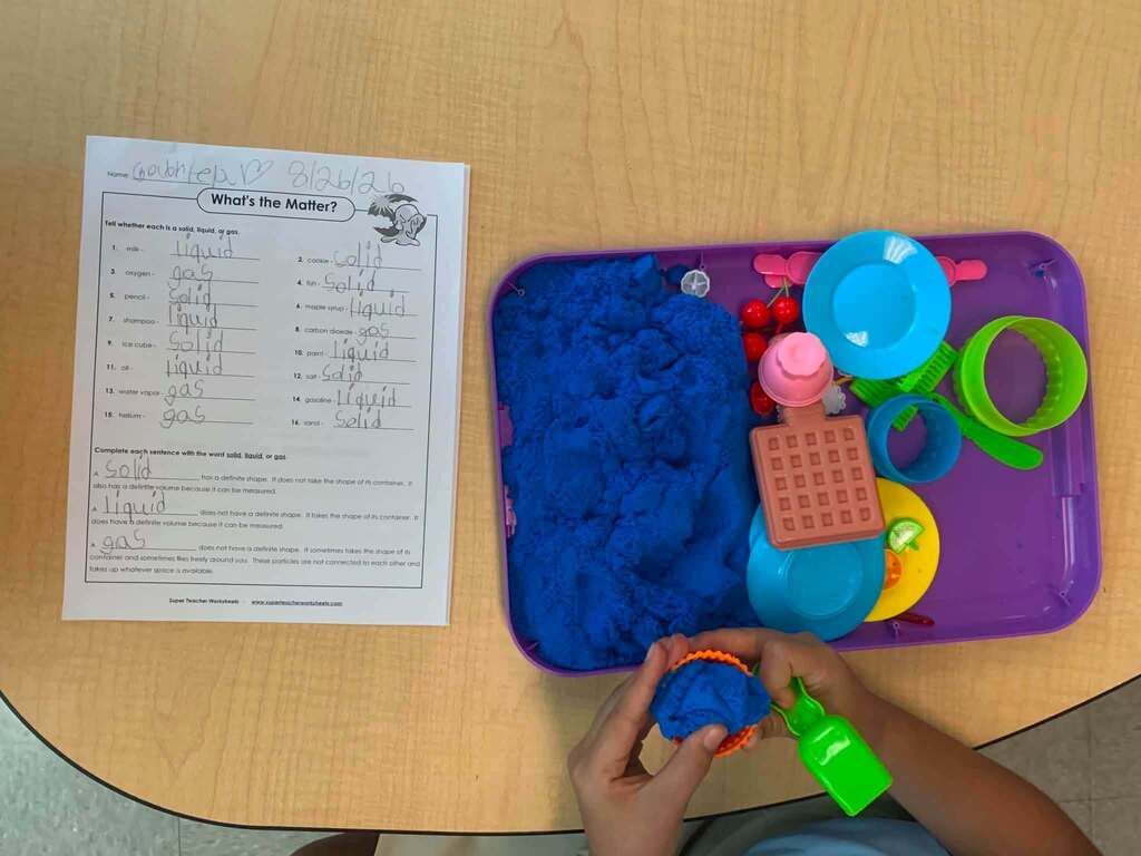 A student is using blue sensory sand with colorful tools beside a worksheet about solids, liquids, and gases.