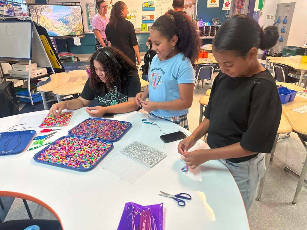 Three students are engaging in a colorful bead craft activity in a school classroom. Other people can be seen in the background.