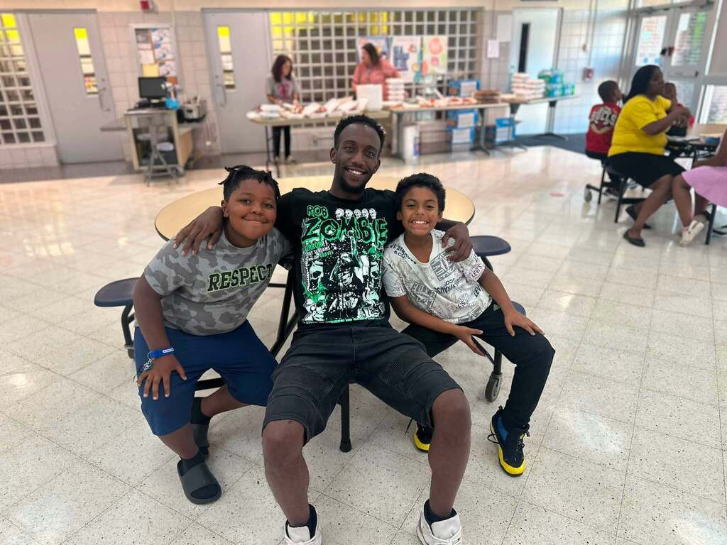 An adult man and two young students are posing together in a school cafeteria. Other people can be seen in the background.