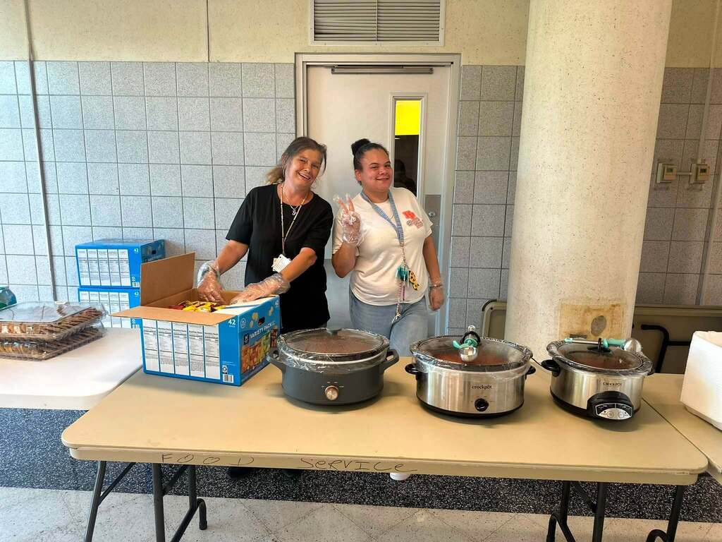 Two district staff members are posing together in a school cafeteria. Food is displayed on the table in front of them.