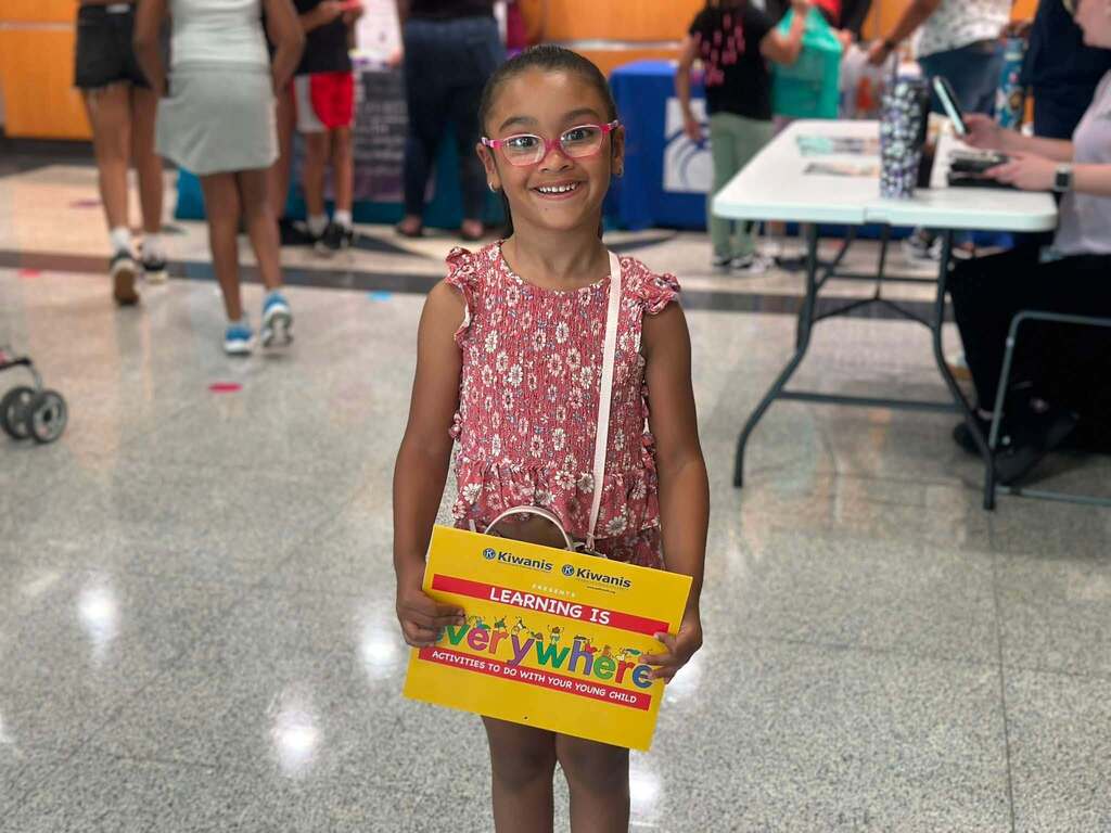 Young student smiling while holding a “Learning Is Everywhere” book at a school event. Other people can be seen in the background.