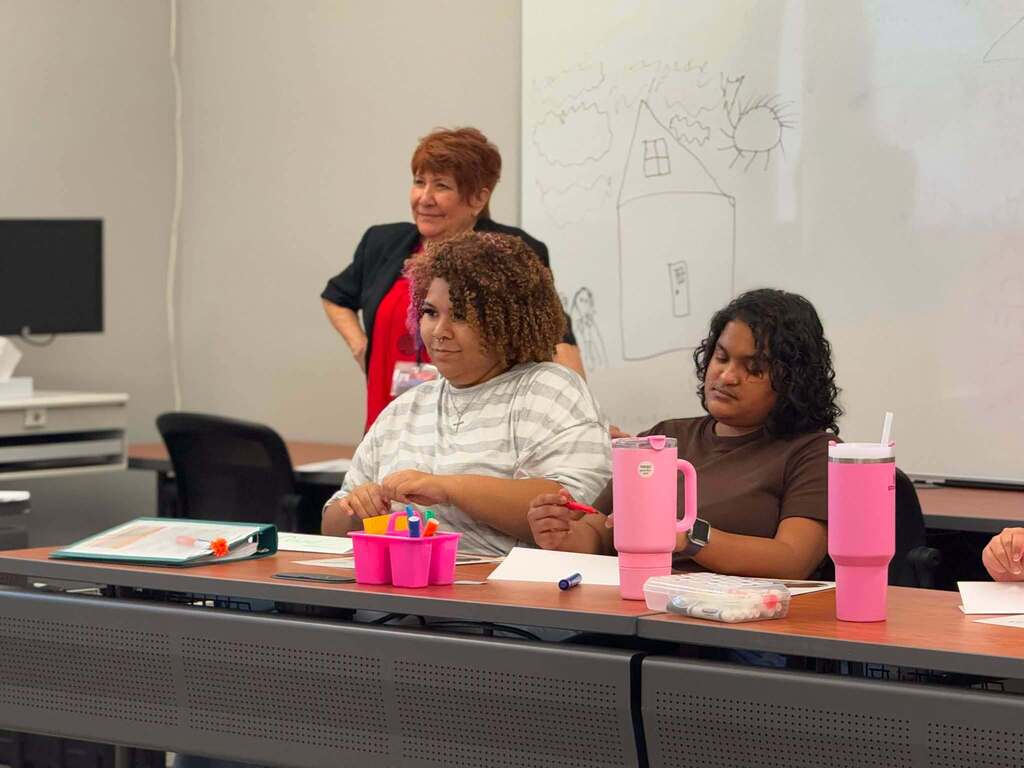 Two students are sitting at a table in a learning lab. An adult woman can be seen in the background.