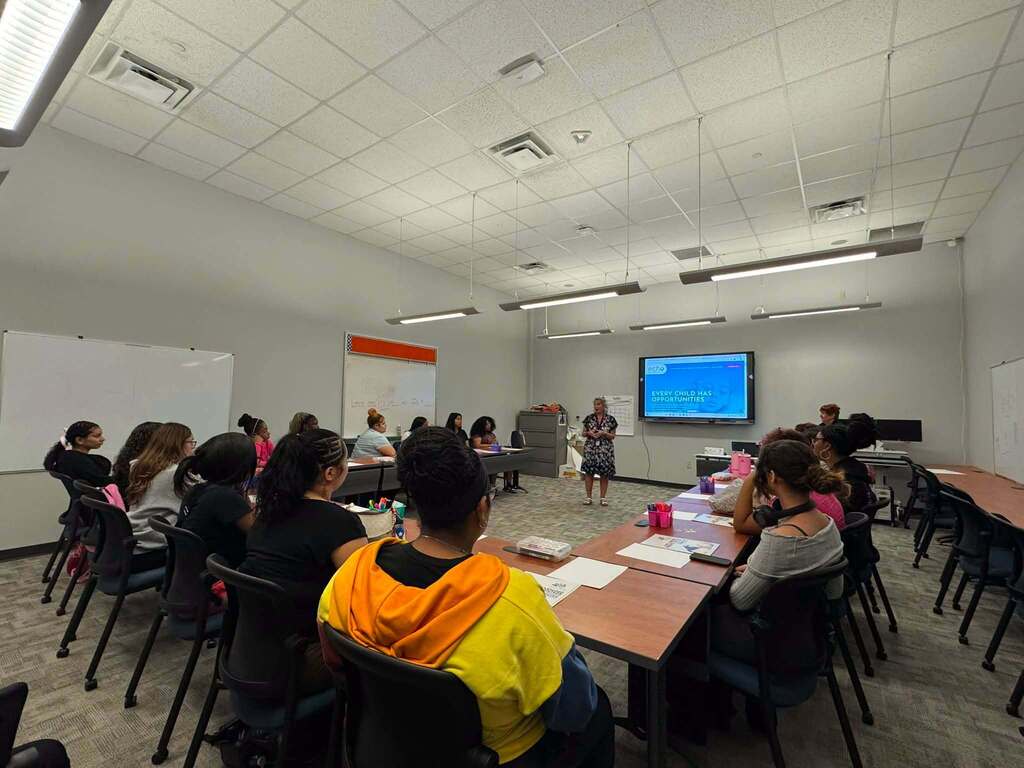A group of students are sitting around tables in a learning lab, listening to an instructor present at the front of the room.