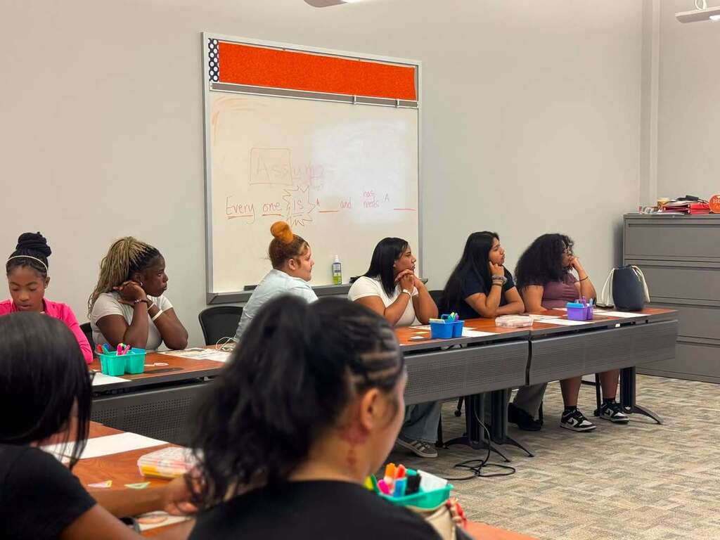 A group of students are sitting around tables in a learning lab, listening to a discussion.