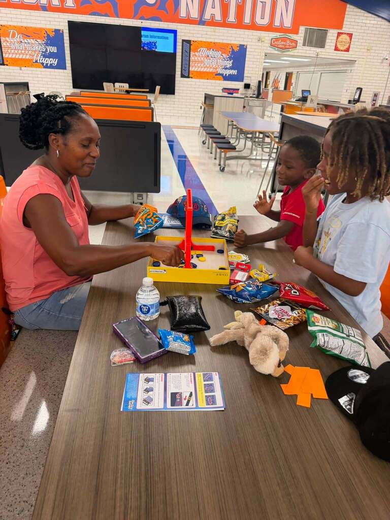 An adult woman and two students are sitting at a table in a school cafeteria playing a board game together.
