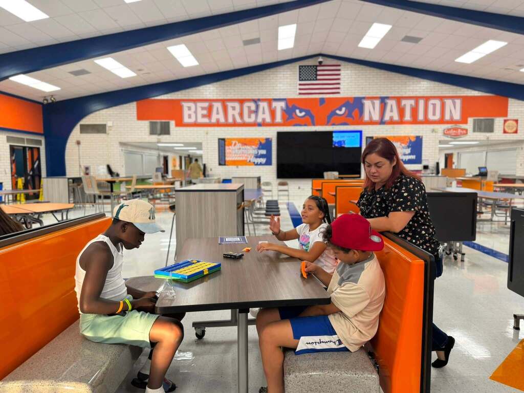 Three students are sitting at a table in a school cafeteria playing a board game together. A district staff member can be seen talking to one of the students.
