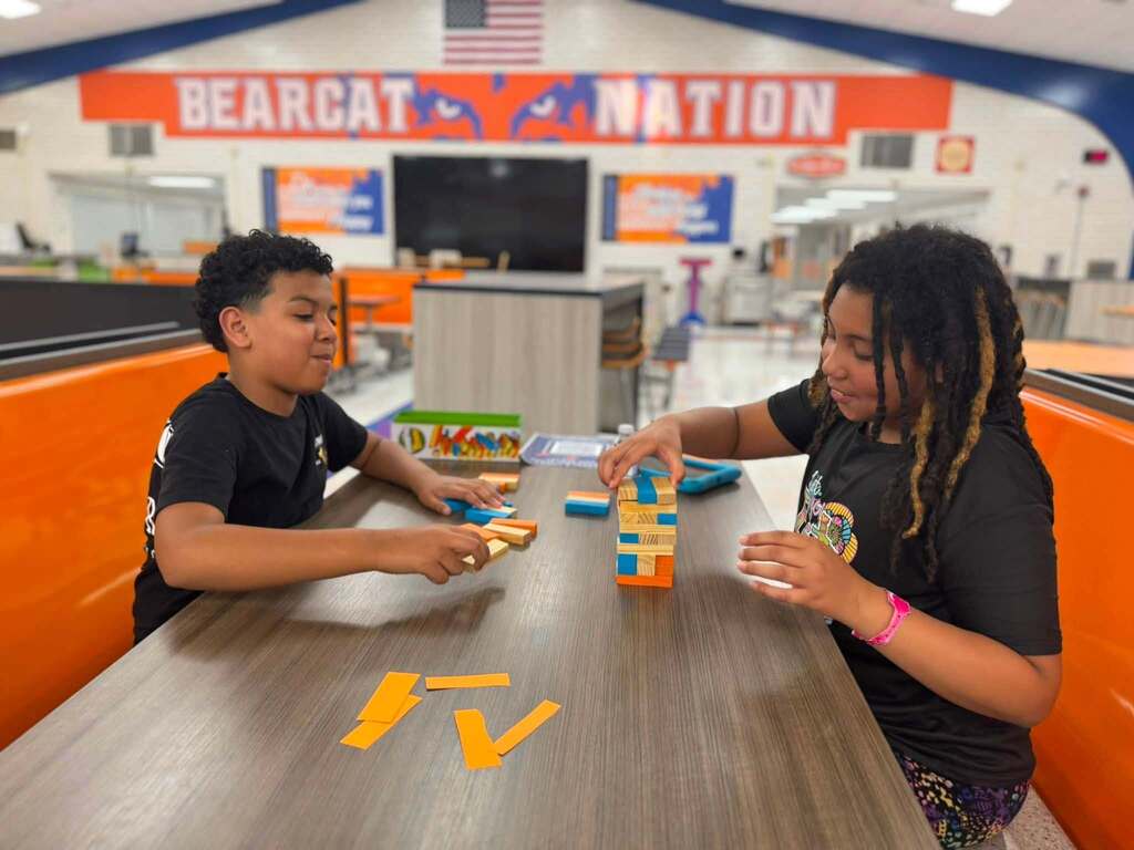 Two students are building a colorful block tower together while sitting at a table in a school cafeteria.