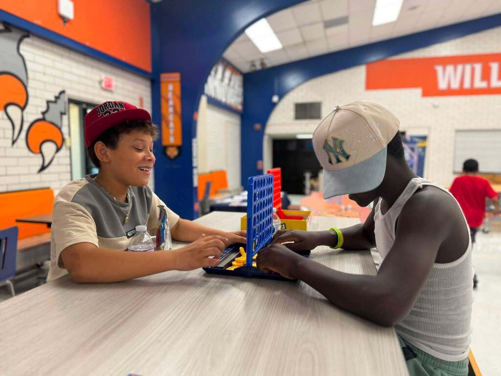 Two students are sitting at a table in a school cafeteria playing Connect Four. Another person can be seen nearby in the background.
