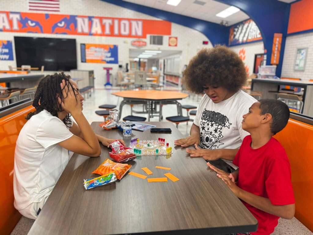 Three students are sitting at a table in a school cafeteria playing a board game together.