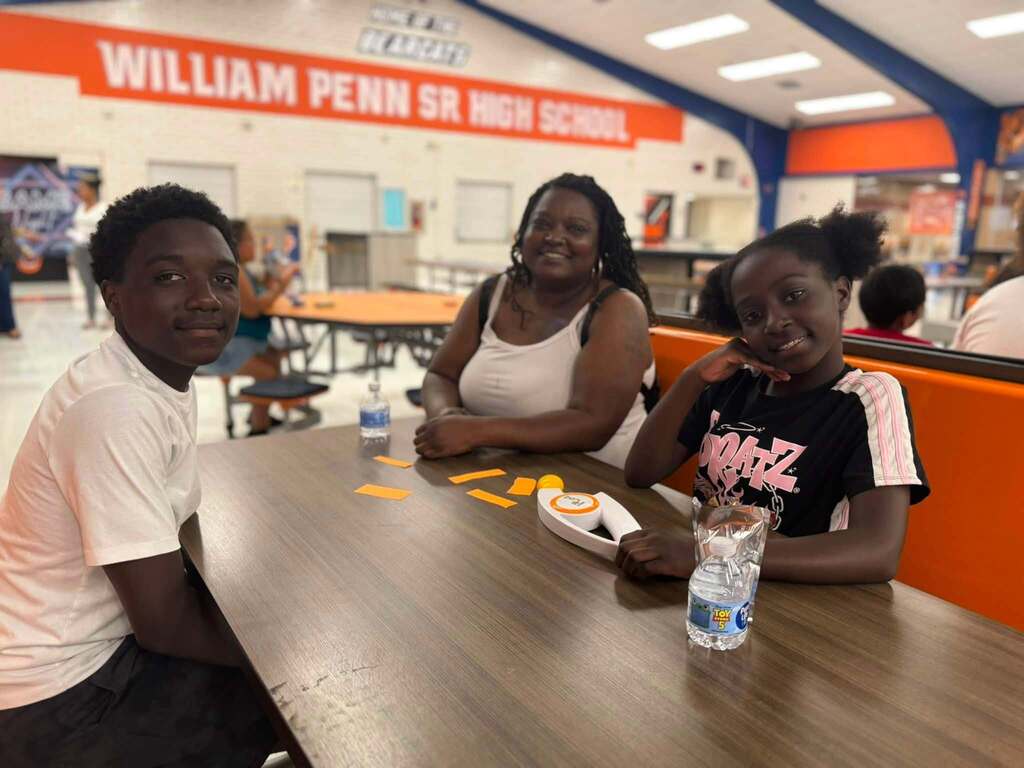 An adult and two students are sitting at a table in school cafeteria playing a game. Other people can be seen nearby in the background.