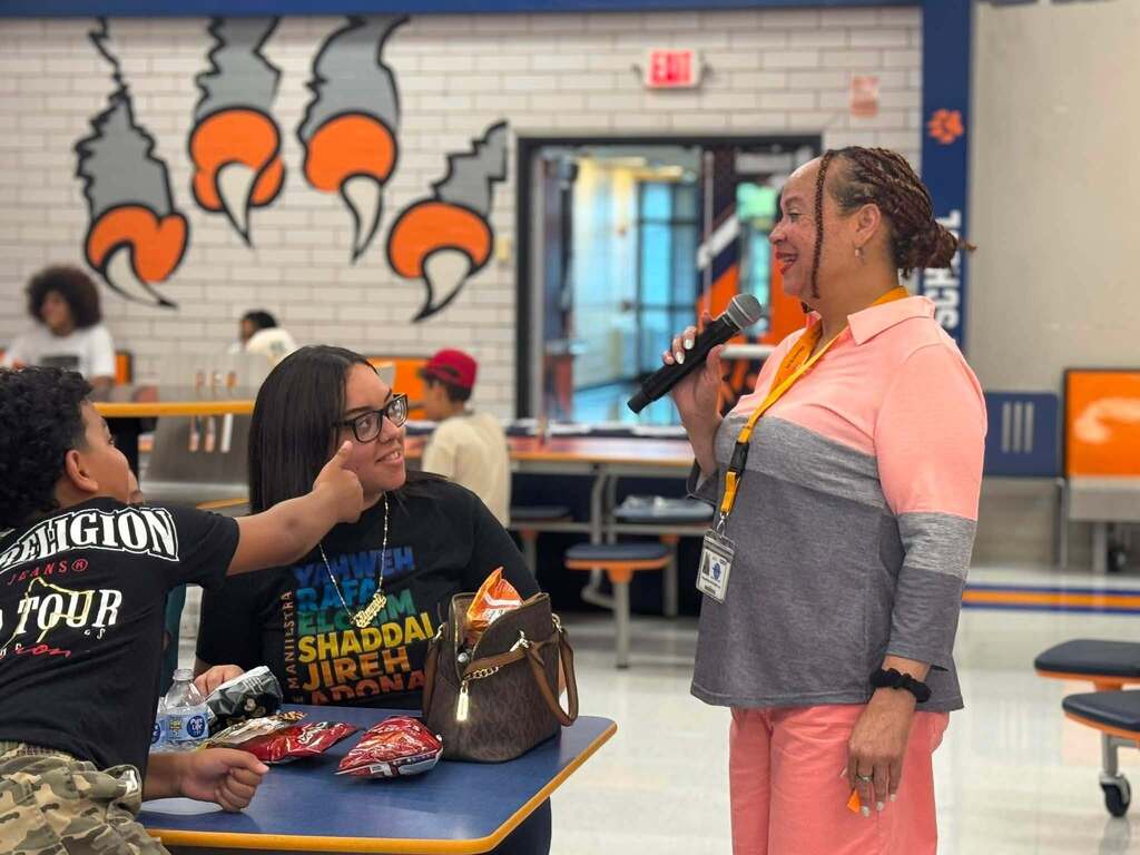 A district staff member is speaking into a microphone while standing in front of an adult woman and a student who are sitting at a table in a school cafeteria. Other people can be seen nearby in the background.